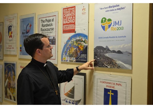 Padre João Chagas diante dos cartazes das JMJ's na sede do PCL em Roma